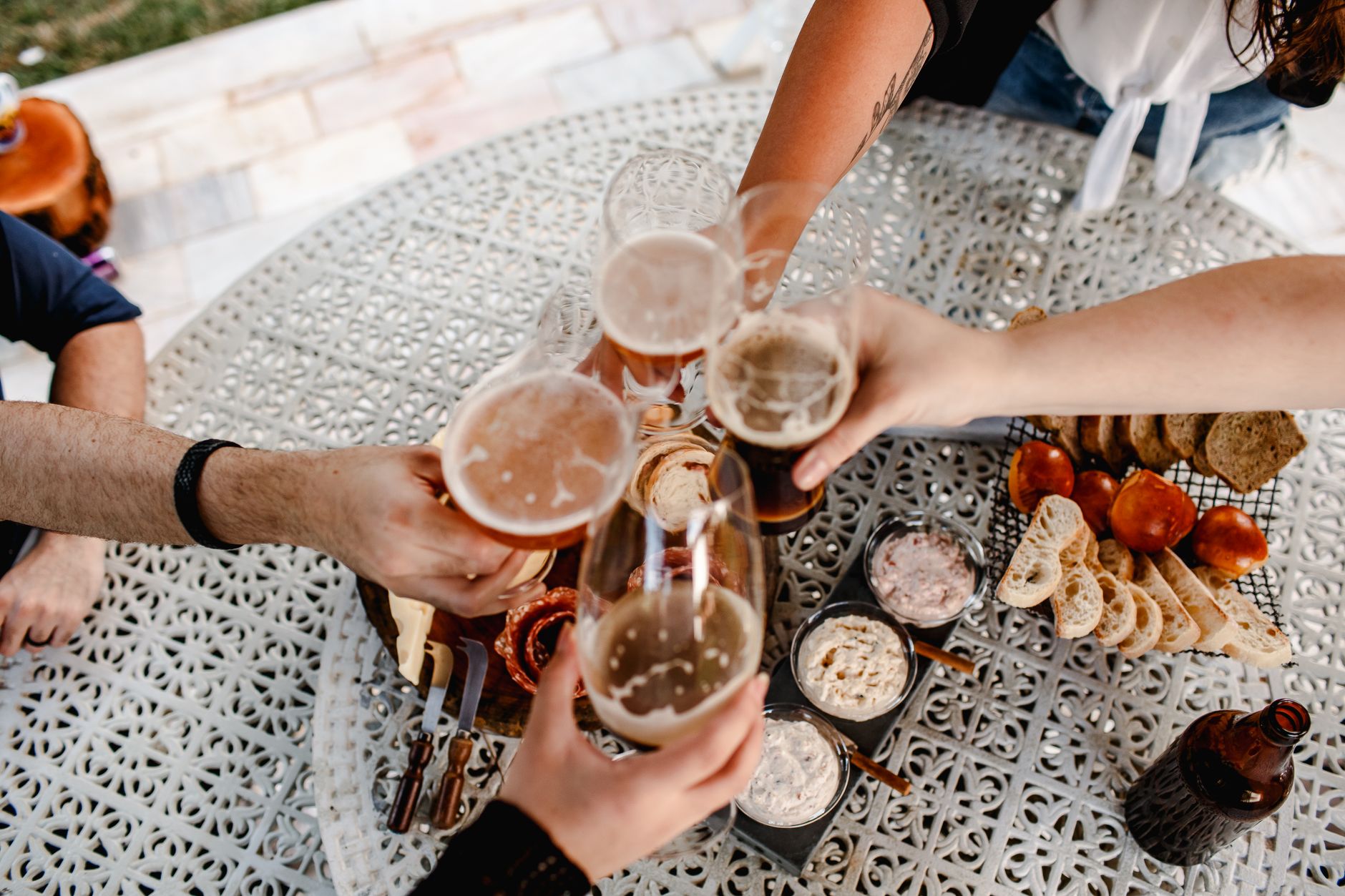 Overhead view of friends clinking beer glasses at a table with charcuterie, enjoying a casual outdoor gathering.